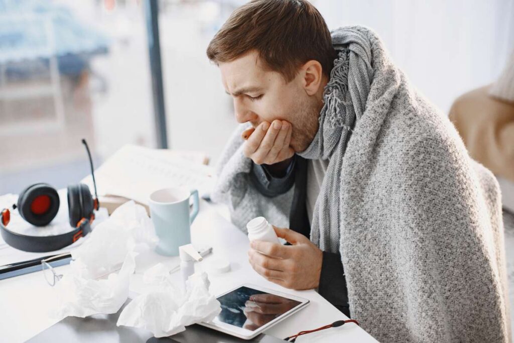 A man experiencing severe symptoms sitting at a desk with tissues and medicine, trying to figure out the difference between a cold, the flu, and COVID