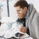 A man experiencing severe symptoms sitting at a desk with tissues and medicine, trying to figure out the difference between a cold, the flu, and COVID