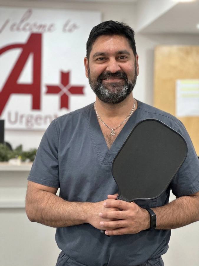 A portrait of Dr. Ajay Jetley, a board-certified Emergency Medicine physician at A+ Urgent Care, standing in clinic holding a pickleball paddle and smiling.