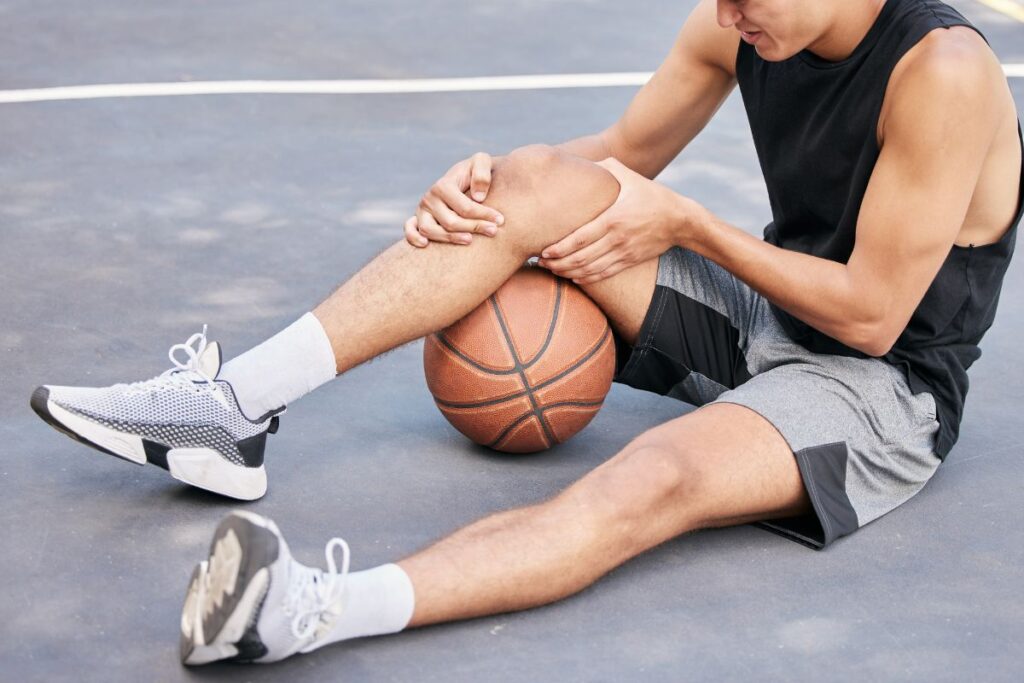 Alt text: A basketball player sitting on an outdoor court holding his injured knee in pain