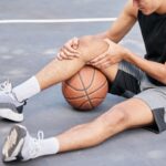 Alt text: A basketball player sitting on an outdoor court holding his injured knee in pain