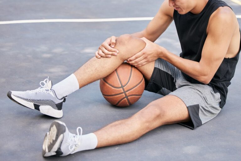 Alt text: A basketball player sitting on an outdoor court holding his injured knee in pain