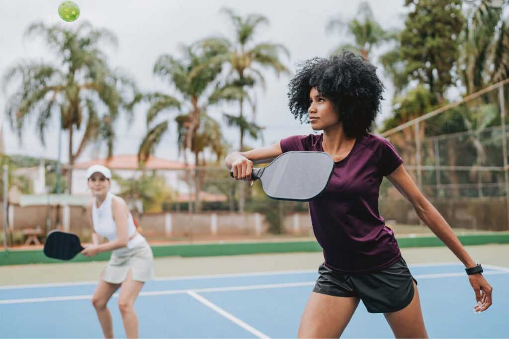A woman plays a pickleball game, holding a paddle mid-swing on an outdoor court. In the foreground, a text overlay reads: "THE RISE OF PICKLEBALL INJURIES: BLACK EYES, TORN CALVES, AND PICKLEBALL ELBOW." A second player is in the blurred background.
