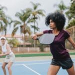 A woman plays a pickleball game, holding a paddle mid-swing on an outdoor court. In the foreground, a text overlay reads: "THE RISE OF PICKLEBALL INJURIES: BLACK EYES, TORN CALVES, AND PICKLEBALL ELBOW." A second player is in the blurred background.