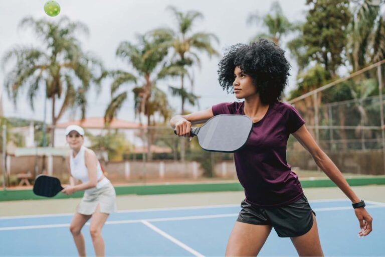 A woman plays a pickleball game, holding a paddle mid-swing on an outdoor court. In the foreground, a text overlay reads: "THE RISE OF PICKLEBALL INJURIES: BLACK EYES, TORN CALVES, AND PICKLEBALL ELBOW." A second player is in the blurred background.